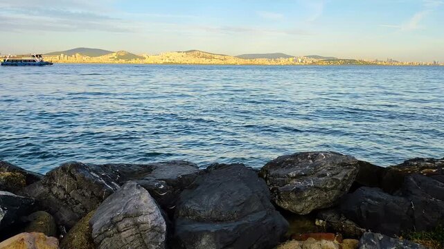 Scenic view of a passenger ferry cruising in the Sea of Marmara with the Istanbul city coastline, rolling hills, and dark seaside rocks in the foreground during daytime.