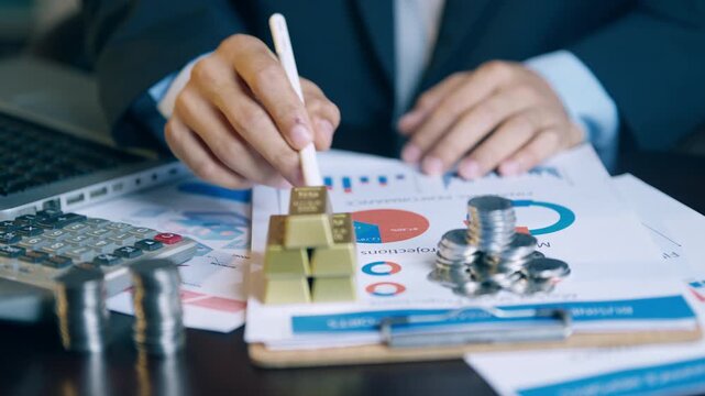 Financial analyst using a pen to check and count gold bars and coins on investment report documents.