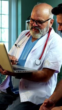 Indian male doctor using laptop while consulting young man patient as experienced physician discusses medical reports in modern Indian clinic OPD healthcare consultation