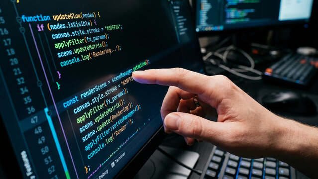 Software developer pointing at computer screen with programming code in a dark office.