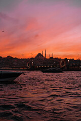 Naklejka premium Yachts and bridge silhouettes below a blazing orange sunset, with Istanbul's skyline and mosque domes glowing beyond the water.