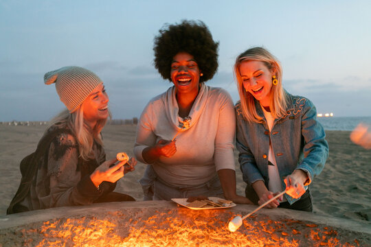 Three women enjoying a beach bonfire, roasting marshmallows together.