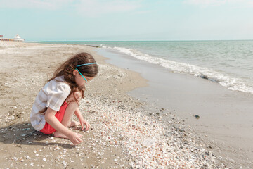Fototapeta premium Young girl crouching on a sandy beach covered in white shells, playing with pebbles near the shoreline with ocean waves in the background.