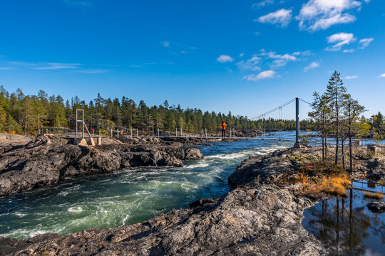 Suspension footbridge crossing the rapids of Trollforsen in northern Sweden, surrounded by rugged rocks, flowing water and boreal forest under a clear blue sky.
