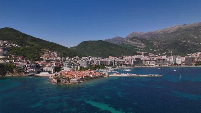 Budva Old Town aerial view over Adriatic Sea with marina and red roofs set against verdant hills and Dinaric Alps. Drone perspective captures coastal layout, hotels, promenades and clear water, drone