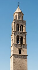 Obraz premium An ancient stone bell tower rises toward a clear blue sky, showcasing intricate masonry, arched windows, and a classic weathered texture, chapel, pinnacle, spire