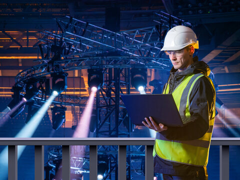 Event production. Stage technician checks lighting truss and concert equipment with laptop during arena show setup.