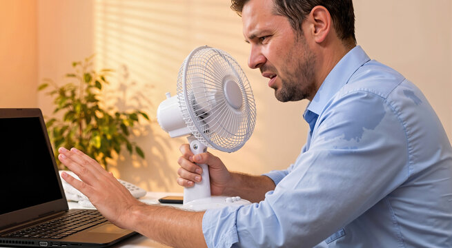 Distressed sweaty man holding a small fan to his face while working at a desk. Overheated male employee with a wet blue shirt looking at a laptop during a severe summer heatwave