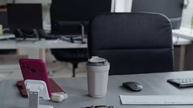 Camera panning over empty office desk with smartphone on stand, glasses, coffee cup, and computer monitor. Video suitable for corporate presentations and remote work marketing campaigns