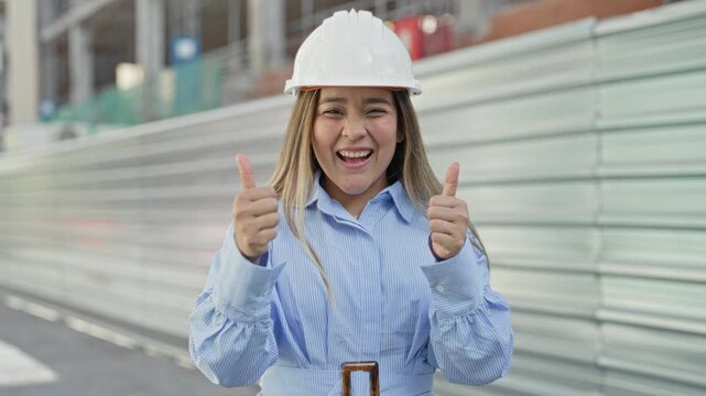 Woman wearing white hardhat giving two thumbs up while smiling in front of metal hoarding at building site; confidence.