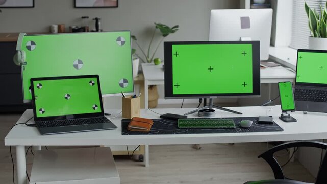 Multiple computer monitors, laptop, and smartphone displaying green screens with tracking markers sitting on office desk. Ideal for showcasing software interfaces via screen replacement