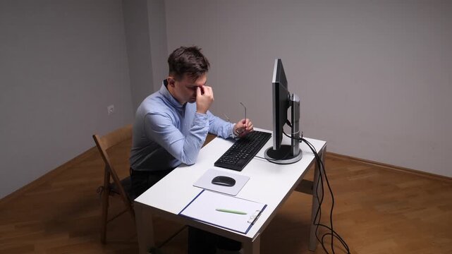Overworked man experiencing headache and eye strain at the office desk, holding his glasses and feeling the pressure of long hours working on the computer