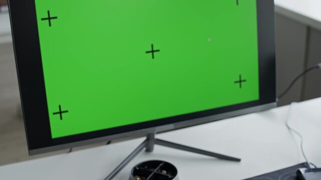 Computer monitor displaying green screen with tracking markers standing on office desk next to keyboard and mouse. Clip serves as mockup for showcasing software development or digital workspace