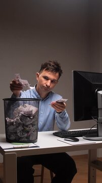 Vertical video disappointed businessman sitting at an office desk, throwing crumpled paper currency into a waste bin, depicting concepts of financial loss, waste, and inflation