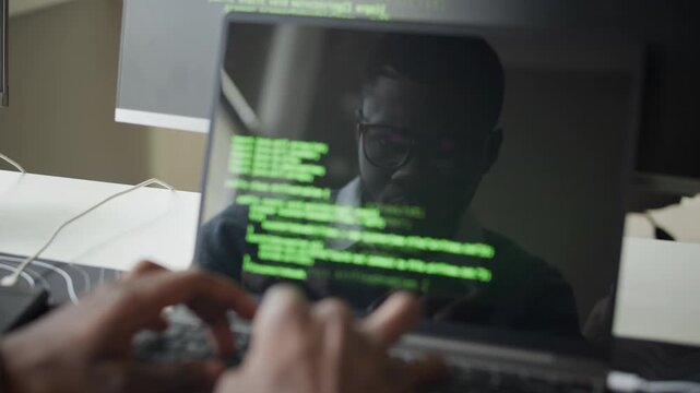 Close up on hands of African American young adult man typing code on laptop with green programming lines reflected on screen. Clip for software development, IT security, and AI technology commercials