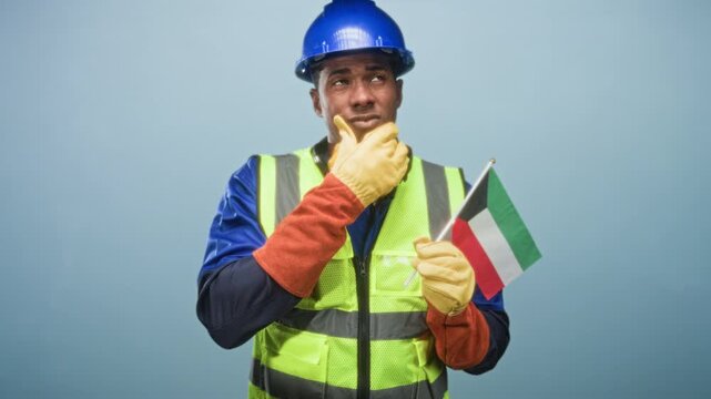 Man construction worker wearing hardhat reflective vest and gloves holding kuwait flag with hand on chin in studio; pride duty.