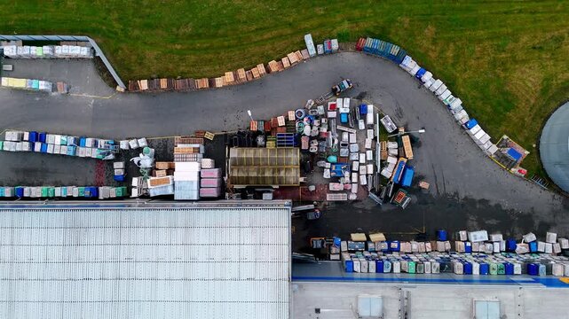 Forklift working in an organized industrial warehouse yard, top down aerial video featuring rows of pallets, construction materials, a curved concrete driveway, building roof, and green grass.