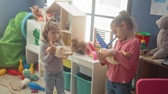 Two girls cradling baby dolls and holding them with both hands in an indoor playroom storage area inside a building surrounded by shelves, toys and bins; tenderness friendship play.