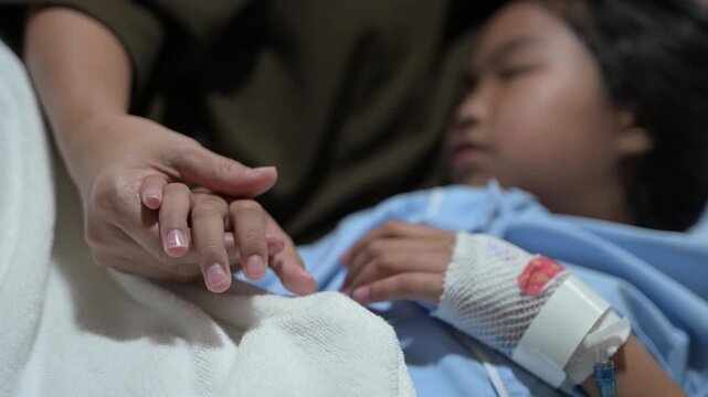 Caring mother holding hand and touching forehead of sick asian little girl patient resting on bed in hospital room. Healthcare, family love, medical recovery and pediatric support concept.