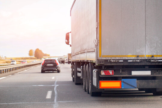 Rear view of large white semi-trailer truck traveling on multilane highway during daytime, other vehicles visible in background, sun glare present