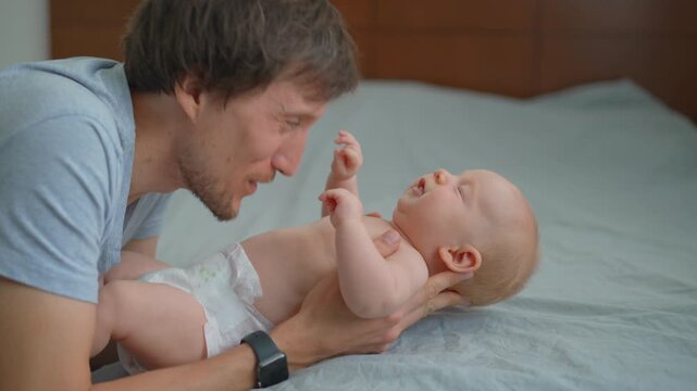 Father kissing cheeks and belly of his three month old baby son on bed.
