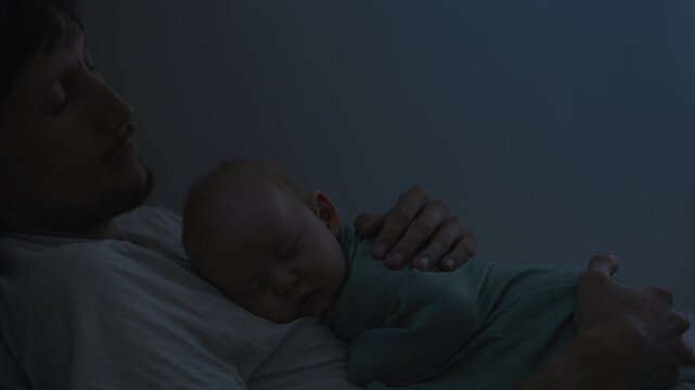 Swaddled baby son sleeping on father's belly at night.