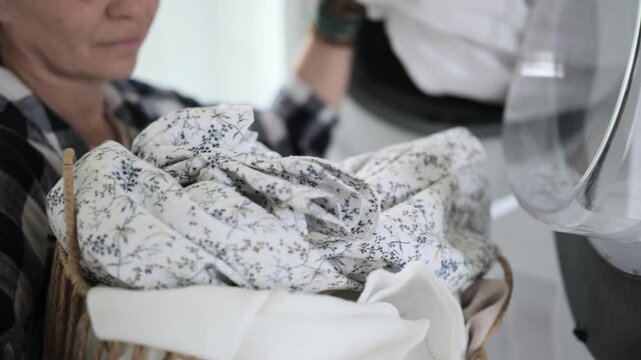 Woman loads patterned fabrics from woven basket into front-load washing machine.