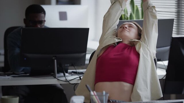 Vertical medium shot of young adult Caucasian woman stretching arms at office desk, then resuming typing on computer. Footage perfect for corporate wellness and software development campaigns