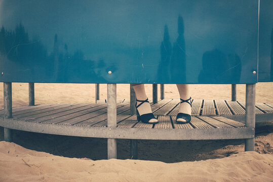 Anonymous woman standing inside a blue beach shelter, visible only by her legs and vintage shoes. Minimalist seaside scene with mystery, nostalgia and a calm summer mood.