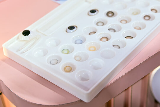 An overhead angled shot of a white plastic display tray containing numerous round slots filled with different colored contact lenses and realistic artificial glass eyeballs