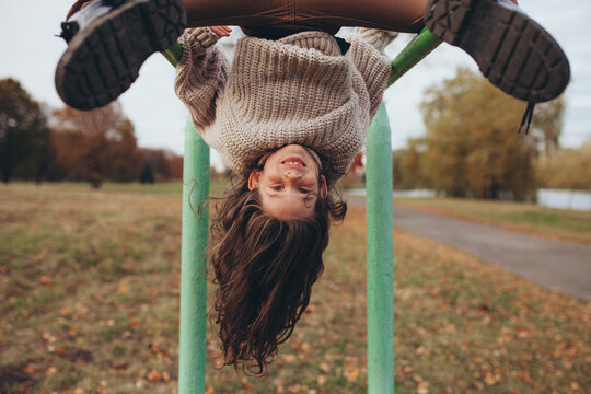 Joyful young girl hanging upside down from metal bar in autumn park. Playful and carefree childhood, happiness, curiosity, and energy.