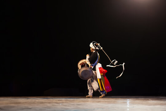 Traditional Korean pungmul performer playing janggu drum with spinning sangmo ribbon on a dark stage during a cultural performance.
