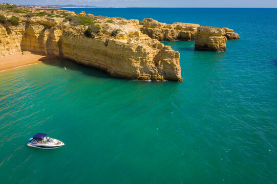 Aerial view of the golden limestone cliffs and crystal clear turquoise waters with people in a small motorboat near a secluded beach in Albufeira, Faro District, Portugal.