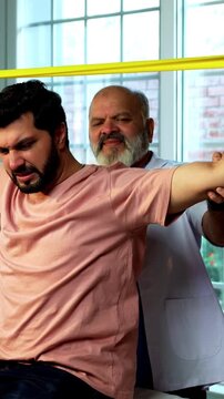 Indian physiotherapist teaching patient resistance band exercises for shoulder and tricep as senior male therapist assists strength training on clinic bed inside modern Indian clinic