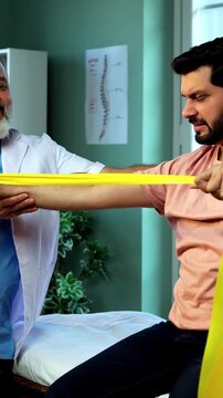 Indian physiotherapist teaching patient resistance band exercises for shoulder and tricep as senior male therapist assists strength training on clinic bed inside modern Indian clinic