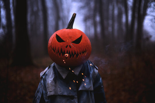 Close up portrait of a sinister pumpkin headed figure with a smoking cigar in the mouth in a dark forest at dusk. Creepy Halloween character with sparks and dramatic lighting creating a haunting mood
