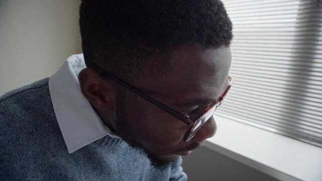 Tilt up shot of young adult Black man typing on keyboard, taking off glasses, and rubbing eyes from exhaustion. Perfect for commercial use depicting digital fatigue, burnout, or remote work stress