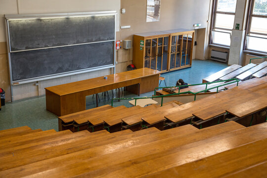 High angle view of a student sitting alone in a large university lecture hall with rows of wooden desks, facing a massive double blackboard and a long wooden professor desk in an old classroom.