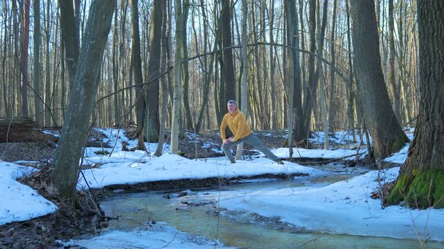 Man stretching legs outdoors in snowy spring forest beside flowing stream, flexibility exercise supporting mobility balance and healthy active lifestyle