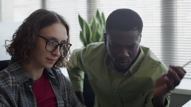 Pull out shot of African American young adult man and Caucasian young adult woman discussing project on computer in office. For tech startup promotions, software development ads, or teamwork concepts