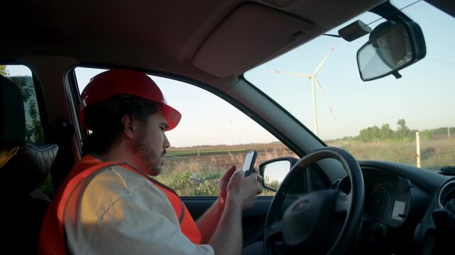 Engine engineer seated inside vehicle performs field inspection using smartphone capturing defect photos checklists coordinates status updates and maintenance report while wind turbines visible