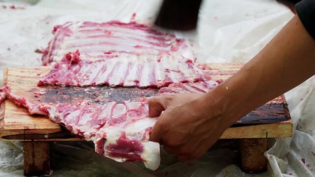 Close-up of unrecognizable people hands cutting fresh raw beef with knives on a board during a traditional communal butcher process or food preparation.