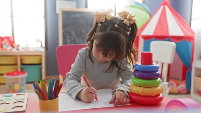 Little girl holding pencil, leaning over paper and writing at a table in a playroom building with stacking rings and crayons; creativity calm learning.