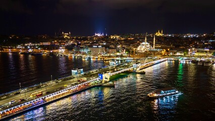 Fototapeta premium Istanbul, Turkey - June 13, 2026: Aerial drone view of Galata Bridge at night, featuring the Golden Horn, bridge lights, waterfront buildings, boats, reflections, and panoramic Istanbul night cityscap