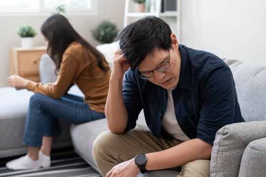 Stressed young Asian man sitting on sofa scratching head after argument with his partner, sad couple having relationship difficulties or fight at home