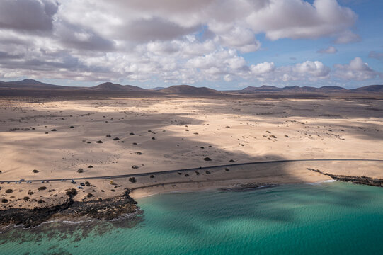 Aerial view of the vast sand dunes of Parque Natural de Corralejo meeting the turquoise ocean and volcanic mountains under a cloudy sky in Corralejo, Canary Islands, Spain.