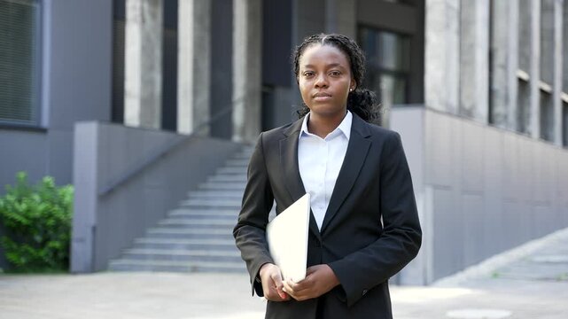 Portrait of a serious black woman standing near a modern office building in the city. A confident professional corporate female manager holds a laptop computer in hands and looks directly into camera.