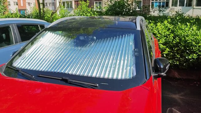 Red car parked outdoors with a reflective sunshade installed on the windshield, showcasing the protective screen against sunlight in a residential area