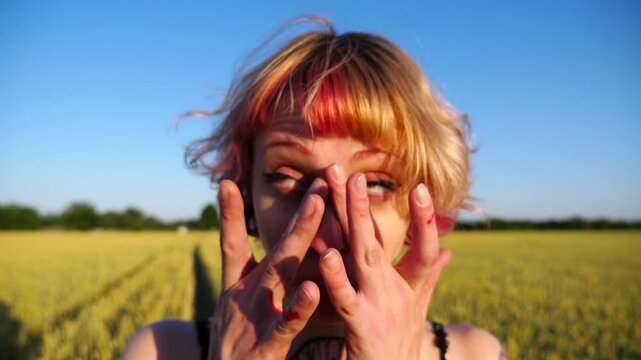 Cheerful punk girl with ear tunnels making funny face while wiping mascara under the eyes with her fingers outdoor. Young hippie woman with pink hair showing grimaces into camera on green meadow