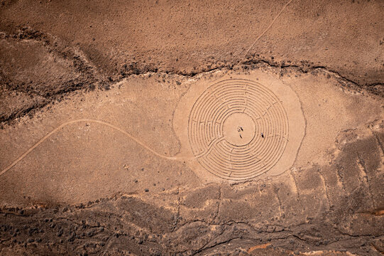 Aerial view of a circular stone labyrinth in the arid desert landscape with people standing in the center and a winding path El Cotillo, Canary Islands, Spain.